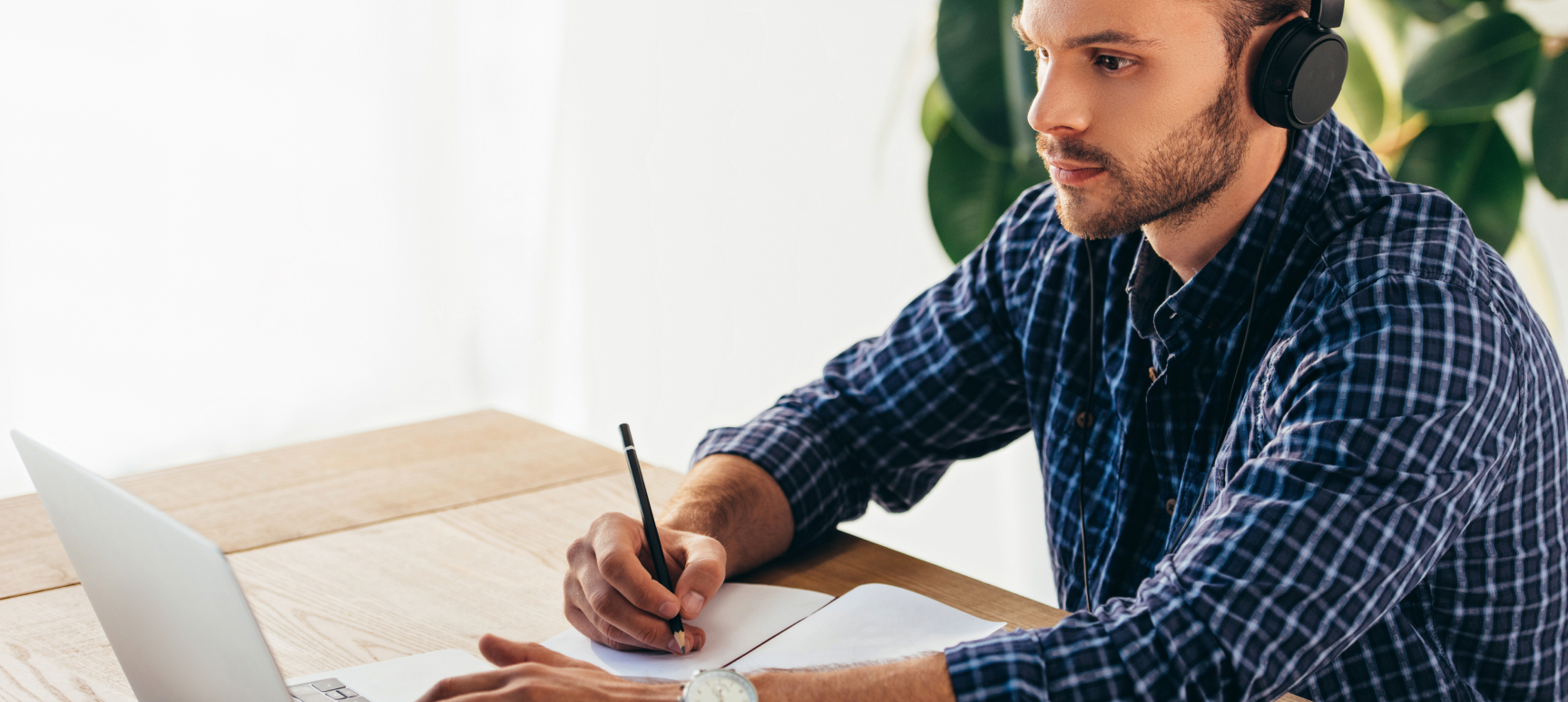 A man sitting at a desk in front of a computer, listening to something through headphones, and writing notes in a journal.